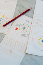 Red colored pencil resting on astrology-style journaling pages with hand-colored hearts, symbols, and circular charts on a marble tabletop