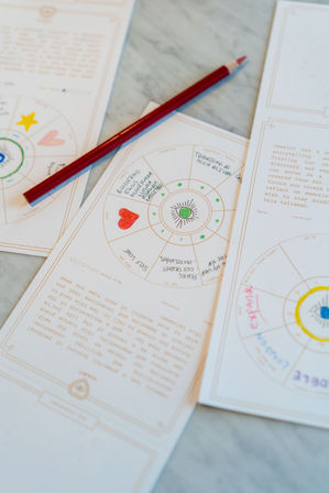Red colored pencil resting on astrology-style journaling pages with hand-colored hearts, symbols, and circular charts on a marble tabletop