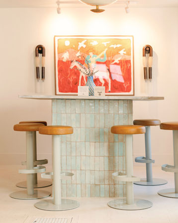 Sunlit boutique café interior with a mint-tiled counter island, caramel leather round stools, blue-striped vase with greenery, and vibrant red wall artwork featuring birds.