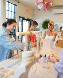 Three women browsing colorful necklaces at a bright boutique jewelry counter with a marble tabletop, necklace display rack, jewelry-making tools, pink pendant light and tropical plant