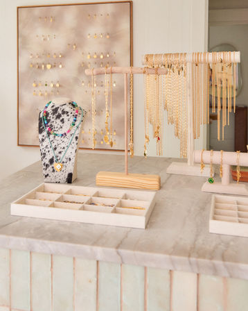 Stylish jewelry display on a marble counter: spotted bust with colorful beaded necklaces and heart pendant, wooden T-stands of gold chains, earring board and compartment trays in a bright boutique setting.