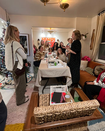 Cozy living-room craft party: a woman leads a group of women and girls gathered around a table with art supplies, snacks, a Christmas tree, and colorful wall art.