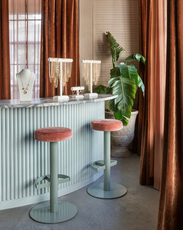 Chic jewelry boutique interior with pale green fluted counter and marble top, two pink velvet bar stools, gold necklace displays on busts, large potted tropical plant, and rust-colored velvet curtains.