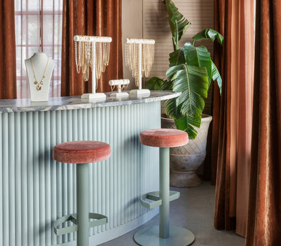 Chic jewelry boutique interior with pale green fluted counter and marble top, two pink velvet bar stools, gold necklace displays on busts, large potted tropical plant, and rust-colored velvet curtains.