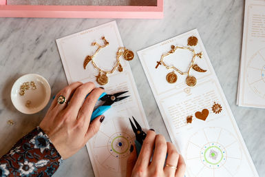 Pair of hands using blue jewelry pliers to assemble handmade gold charm bracelets on printed astrological cards at a marble crafting station, with a small dish of jump rings and loose charms nearby.