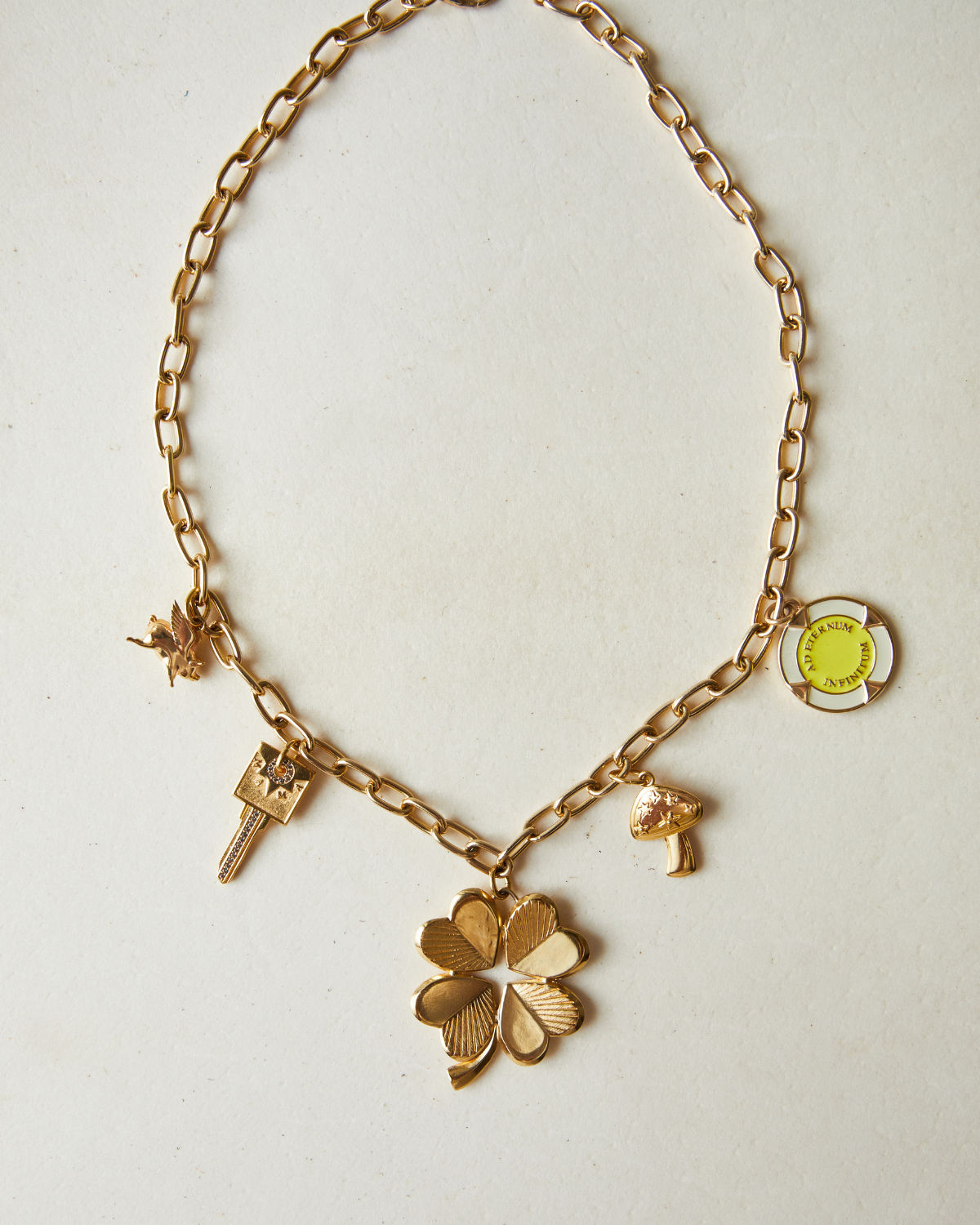 Gold charm necklace flat-lay on off-white background with a large four-leaf clover pendant and dangling key, bird, mushroom charms and yellow enamel round tag