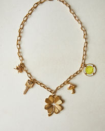 Gold charm necklace flat-lay on off-white background with a large four-leaf clover pendant and dangling key, bird, mushroom charms and yellow enamel round tag