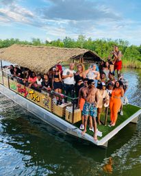 Tiki-style floating party barge on a calm river with a large group of friends in summer attire enjoying drinks and music near a green shoreline