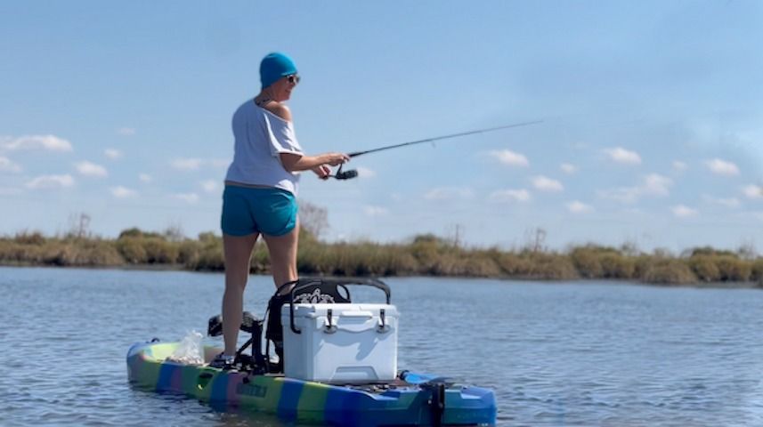 Person standing on a colorful sit-on-top kayak, casting a fishing rod on calm marsh waters under a bright blue sky, with a white cooler and gear on the deck.