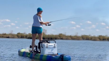 Person standing on a colorful sit-on-top kayak, casting a fishing rod on calm marsh waters under a bright blue sky, with a white cooler and gear on the deck.
