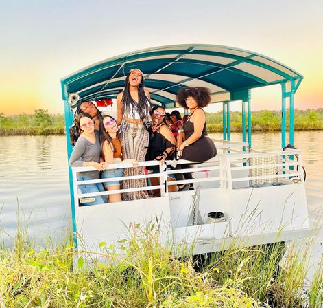 Group of friends laughing and posing on a teal-canopied pontoon boat at sunset over calm marsh wetlands, tall grasses in the foreground — lively outdoor boat ride.