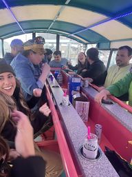 Adults laughing and drinking aboard a covered pedal pub/bar with pink countertops and neon strip lights, canned cocktails in cup holders and party hats, riding through city streets.
