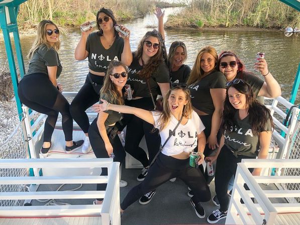 Group of friends celebrating a New Orleans (NOLA) bachelorette on a pontoon boat in a bayou — bride in a white 'NOLA' tee and crew in matching shirts, holding drinks and smiling.