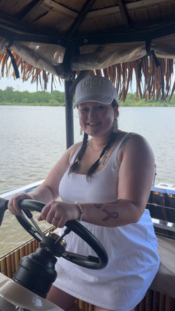 Smiling person in a 'BRIDE' cap steering a thatched-roof pontoon boat on a calm river with green shoreline, wearing a white dress and braided hair