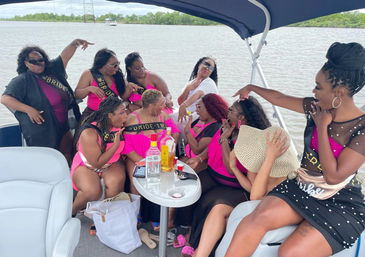Bachelorette boat party on a coastal bay — women in pink outfits and Bride sashes laughing around a table with bottles and cups, water and mangroves in the background.