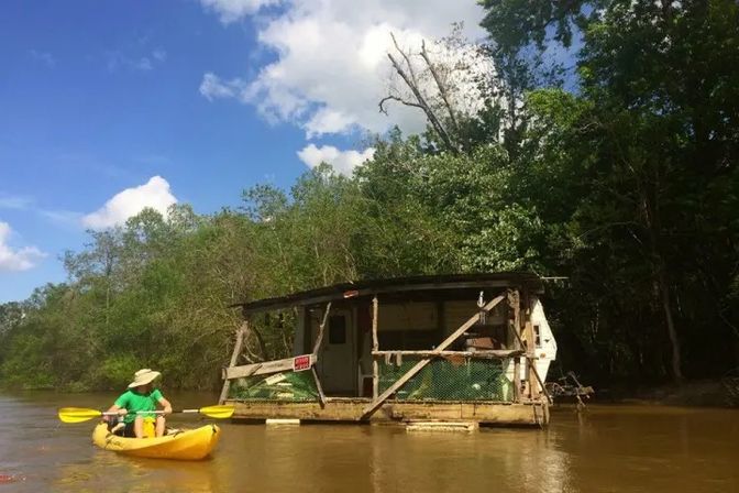Kayaker in a yellow kayak paddling past a rustic floating shack on a muddy river, flanked by dense green trees under a bright blue sky.