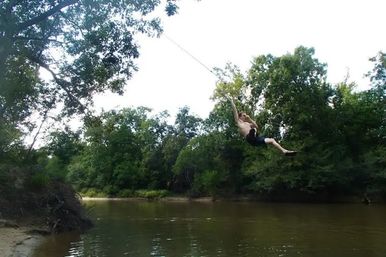 Person midair on a rope swing over a wooded river bend, brown water below, sandy bank and dense green trees on a sunny summer day