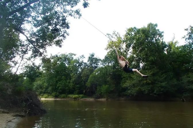 Person midair on a rope swing over a wooded river bend, brown water below, sandy bank and dense green trees on a sunny summer day
