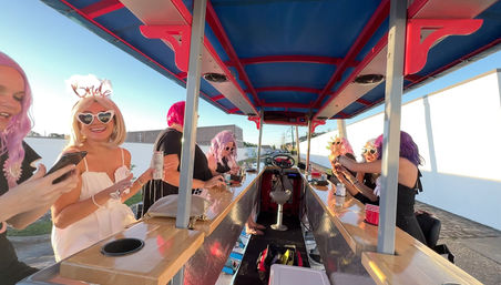 Bachelorette group riding a colorful outdoor pedal bar at sunset — bride wearing a "bride" headband and heart-shaped sunglasses with friends in pink and purple wigs, holding drinks along wooden counters with a steering wheel and sunny street backdrop.