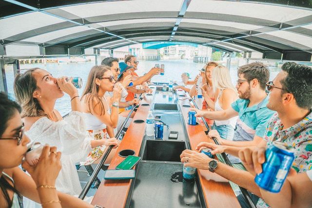 Smiling group on a covered party boat sharing canned drinks around a central bar table, cruising a sunny waterfront canal with bridges and buildings in the background.