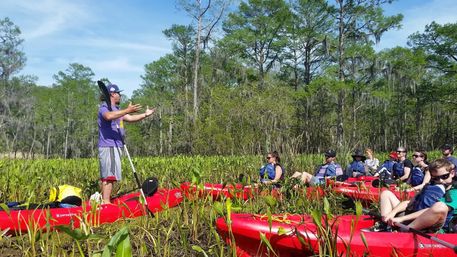 Group of kayakers in bright red kayaks in a cypress swamp with Spanish moss; an instructor stands on a kayak holding a paddle and gesturing while participants listen among green marsh plants under a blue sky.