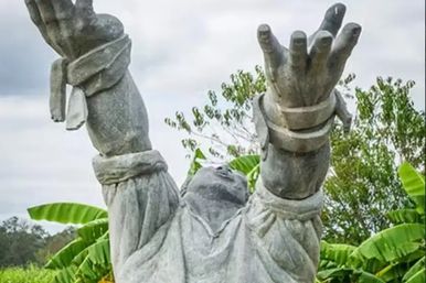 Gray stone statue of a robed figure reaching skyward with raised hands, surrounded by banana leaves and tropical garden greenery under a cloudy sky