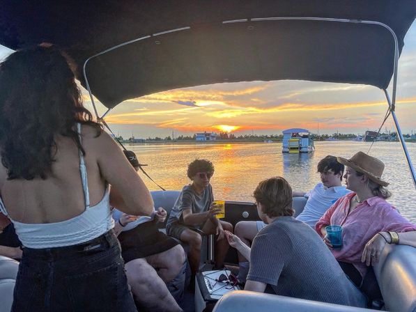 Friends enjoying a relaxed sunset cruise on a pontoon boat, colorful orange-pink sky reflecting on calm water, drinks and conversation under a shaded canopy
