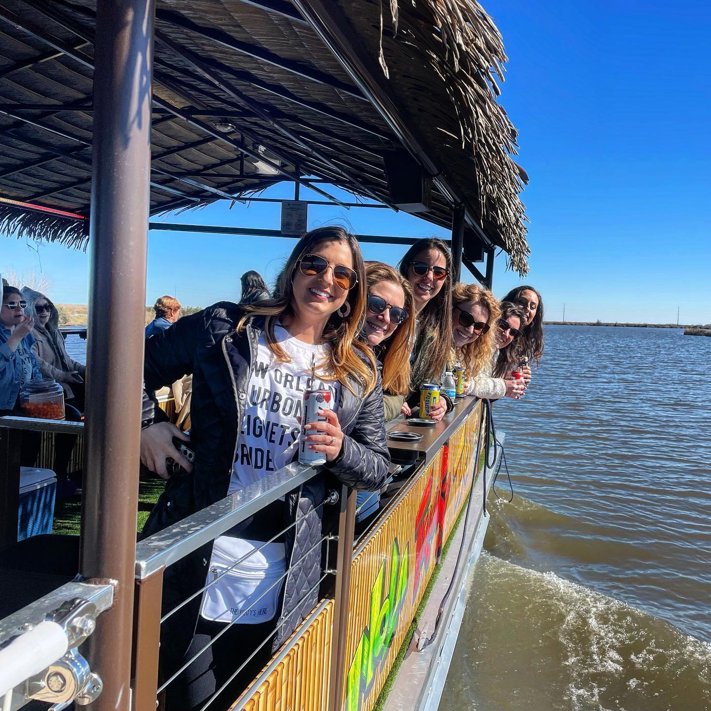 Smiling group of friends leaning over the railing of a tiki-style floating bar, holding canned drinks on a sunny river cruise with blue sky and marshy shoreline in the background.