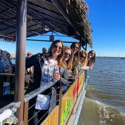Smiling group of friends leaning over the railing of a tiki-style floating bar, holding canned drinks on a sunny river cruise with blue sky and marshy shoreline in the background.
