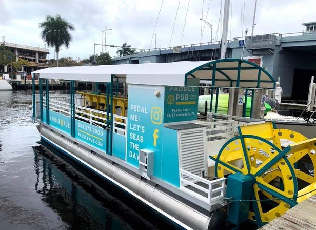 Teal-and-yellow pedal-powered party boat with covered seating and large paddle wheels docked on a Fort Lauderdale canal under a bridge