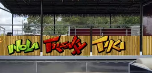 Outdoor covered tiki patio with a bamboo fence featuring bright green, red, and yellow graffiti-style lettering, metal railing and trees visible behind the seating area.
