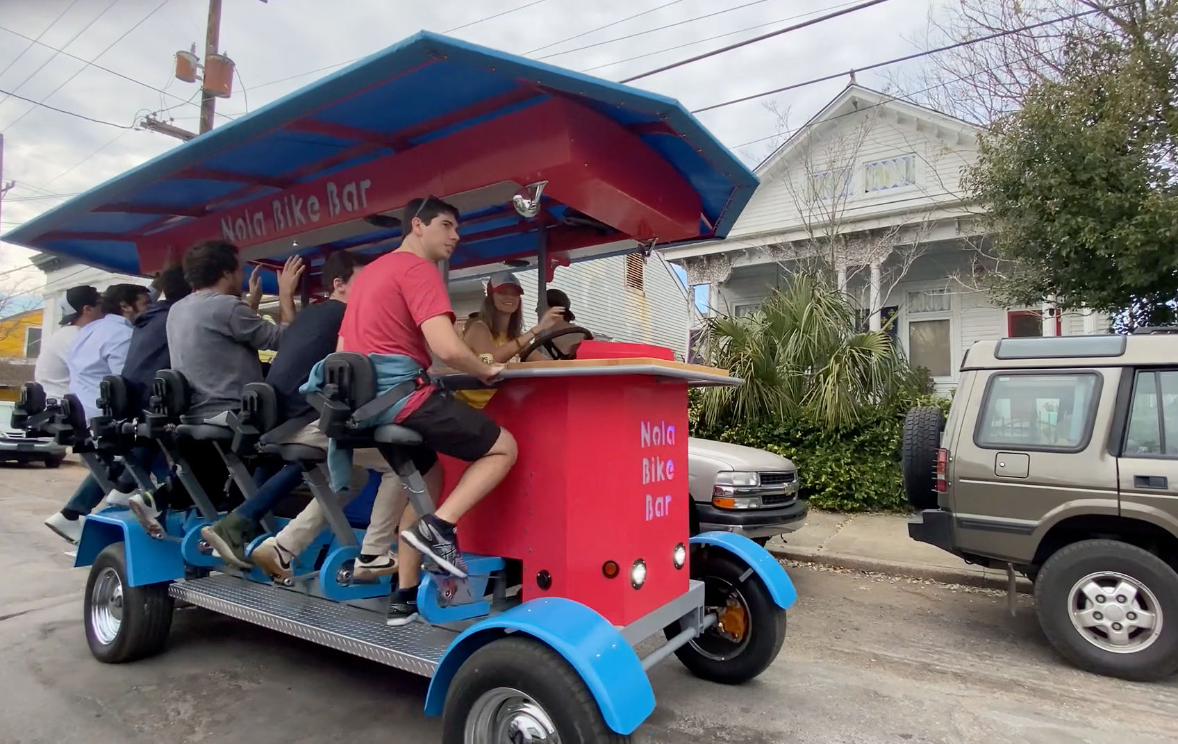 Colorful red-and-blue party pedal bar with a group pedaling along a New Orleans residential street, passengers on bar-style seats pedaling while one person steers.