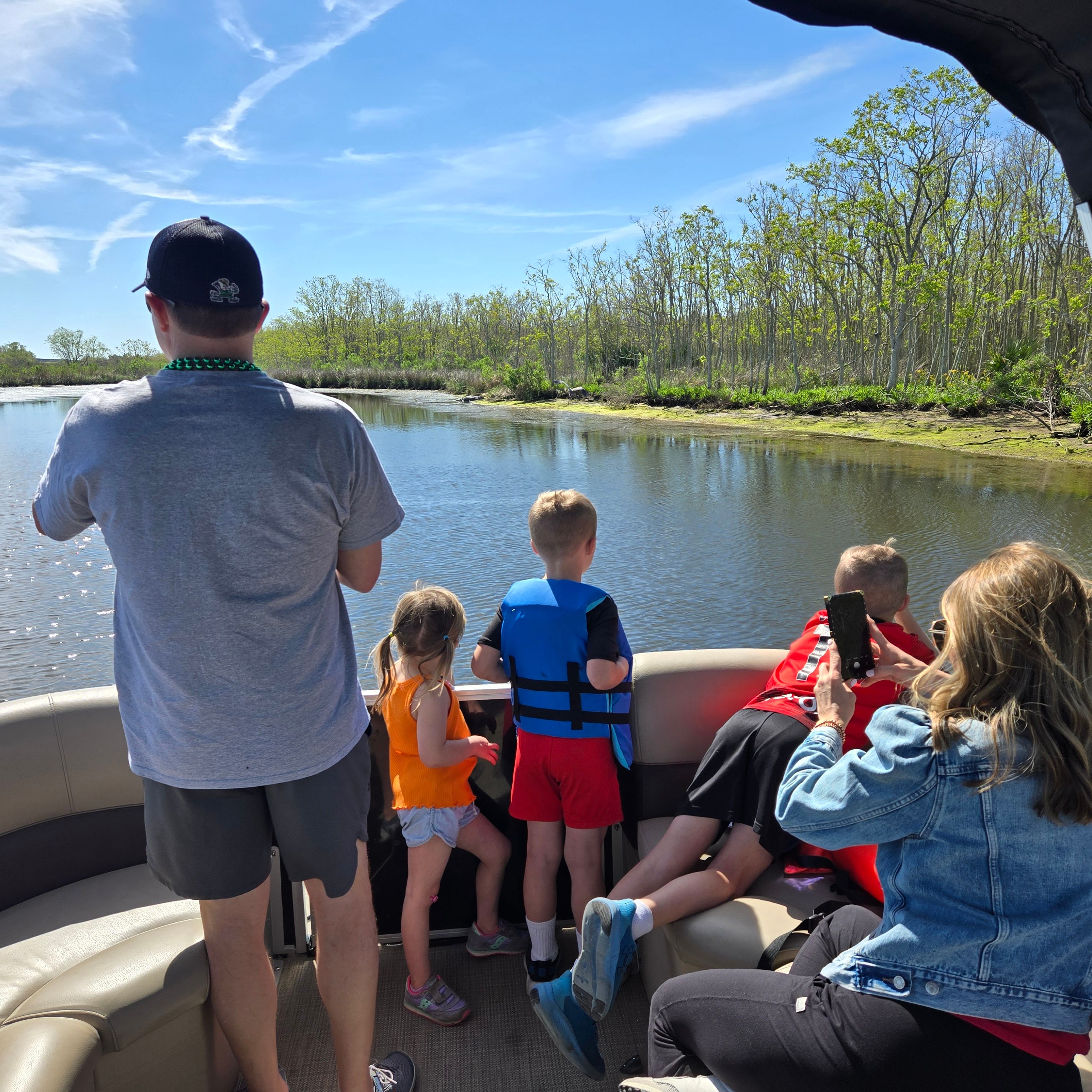 Kids in life jackets peering over the railing of a pontoon boat as adults snap photos on a sunny day, looking out at a calm river bordered by green marshland and trees.