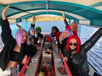 Cheerful group wearing colorful wigs and jackets partying on a covered pontoon boat, raising drinks over a center bar stocked with snacks and beverages, lake and tree-lined shore in the background.