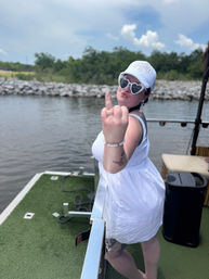 Person in a white 'BRIDE' cap and heart-shaped sunglasses on a pontoon boat at a lake, wearing a white dress and playfully flipping the middle finger at the camera, grassy deck with rocky shoreline and trees in the background.