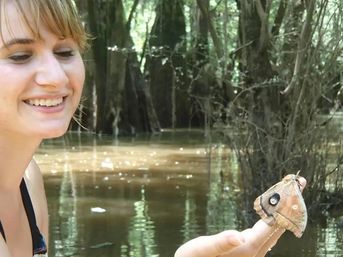 Smiling person holding a large brown moth with a prominent eye-spot on its wing perched on a fingertip in a sunlit swampy forest
