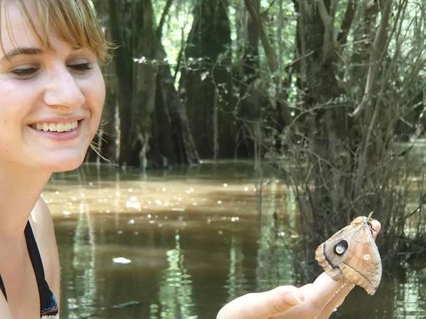 Smiling person holding a large brown moth with a prominent eye-spot on its wing perched on a fingertip in a sunlit swampy forest