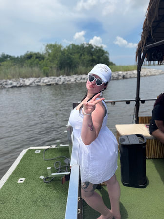 Person in a white sundress and bride cap flashing a peace sign and wearing heart-shaped sunglasses on a pontoon boat deck with a rocky river shoreline and green trees under a partly cloudy sky
