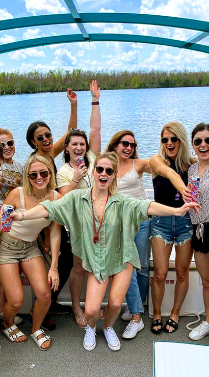 Group of friends cheering on a pontoon boat over a sunny lake, wearing sunglasses and holding colorful drinks — summer boat outing.