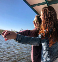 Two friends on a sunny boat at the waterfront, smiling and holding hands out over calm river water — one wearing sunglasses, the other in a denim jacket.