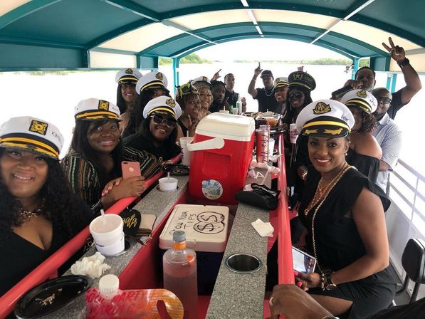 Group of adults wearing captain hats smiling around a long bar with coolers and drinks on a covered pontoon boat during a daytime lake boat party.