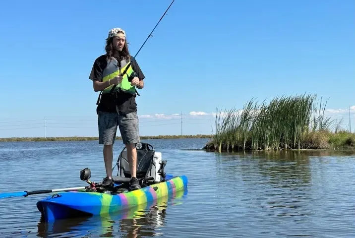 Standing kayaker fishing on a rainbow-striped kayak in calm marsh waters, wearing a neon life vest with reeds and clear blue sky in the background
