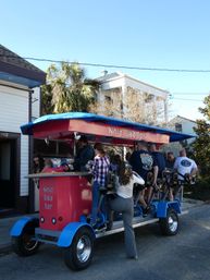 Group pedaling a red-and-blue pedal-powered party bar on a sunny street lined with palm trees and historic houses, lively outdoor tour vibe.
