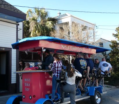 Group pedaling a red-and-blue pedal-powered party bar on a sunny street lined with palm trees and historic houses, lively outdoor tour vibe.