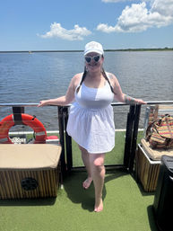 Person in white sundress and heart-shaped sunglasses standing barefoot on a sunny boat deck, overlooking a calm river and blue sky with a life ring visible.