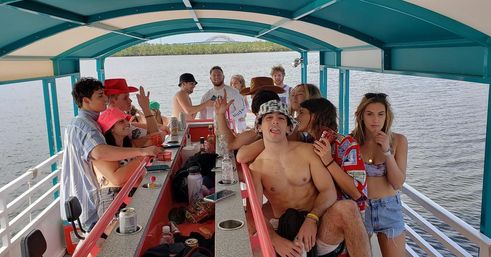 Group of friends having a lively boat party on a covered pontoon bar, holding drinks and smiling on a calm river with a bridge and tree-lined shore in the background