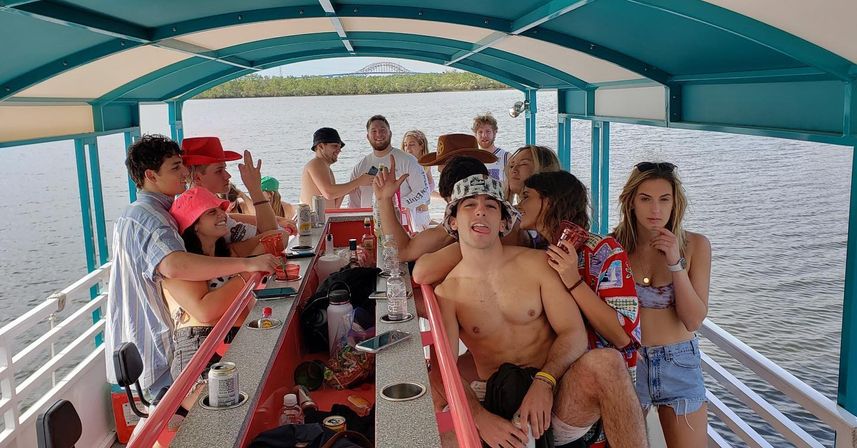 Group of friends having a lively boat party on a covered pontoon bar, holding drinks and smiling on a calm river with a bridge and tree-lined shore in the background