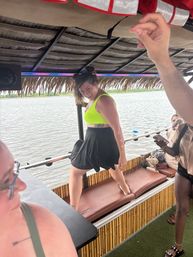 Smiling woman in neon green top and black skirt dancing on a cushioned bench aboard a tiki-style party boat on a calm lake with friends nearby.