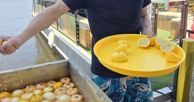 Person on a waterfront dock squeezing a lemon over a metal tub filled with floating onions and lemons, holding a yellow tray with lemon halves.