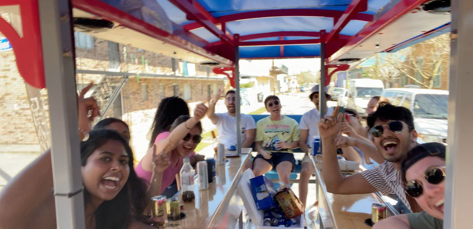 Adults laughing and cheering aboard a red-canopied party bike (pedal pub) with canned drinks on the central bar, rolling down a sunny downtown street.